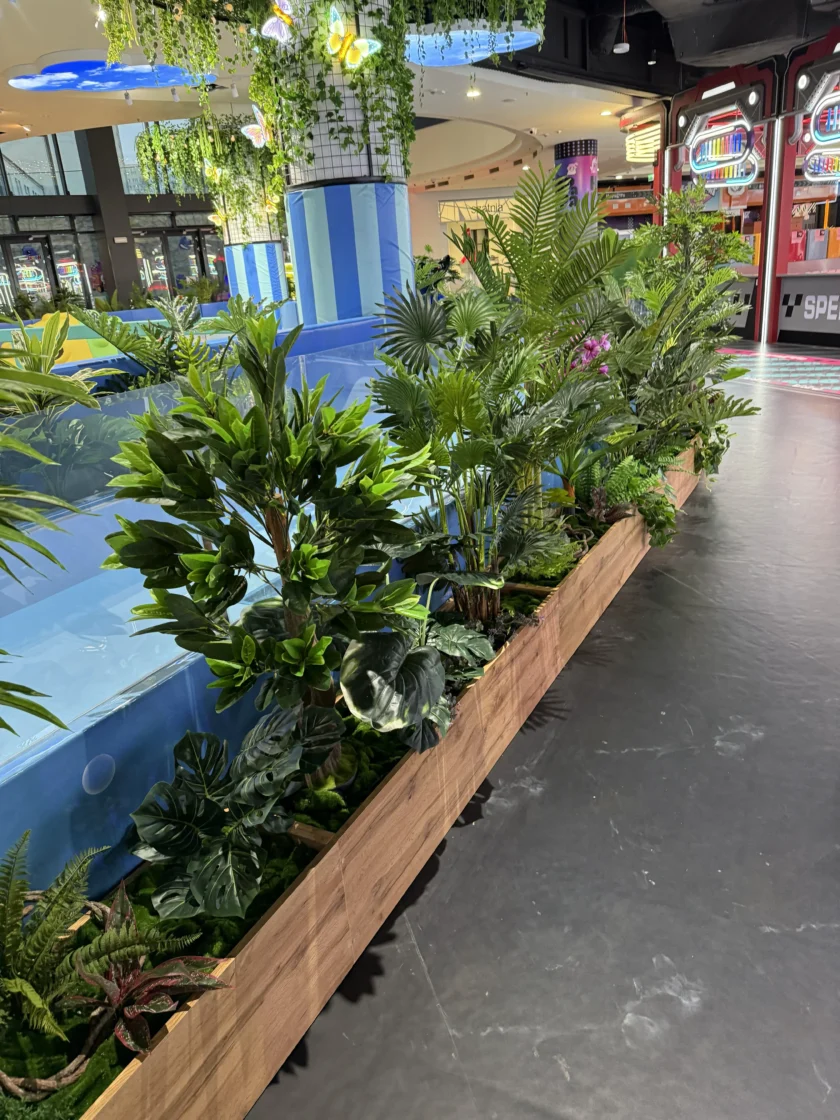 Rząd różnych zielonych roślin doniczkowych w drewnianej donicy obok niebieskiej konstrukcji wewnętrznej w jasno oświetlonym miejscu rozrywki.