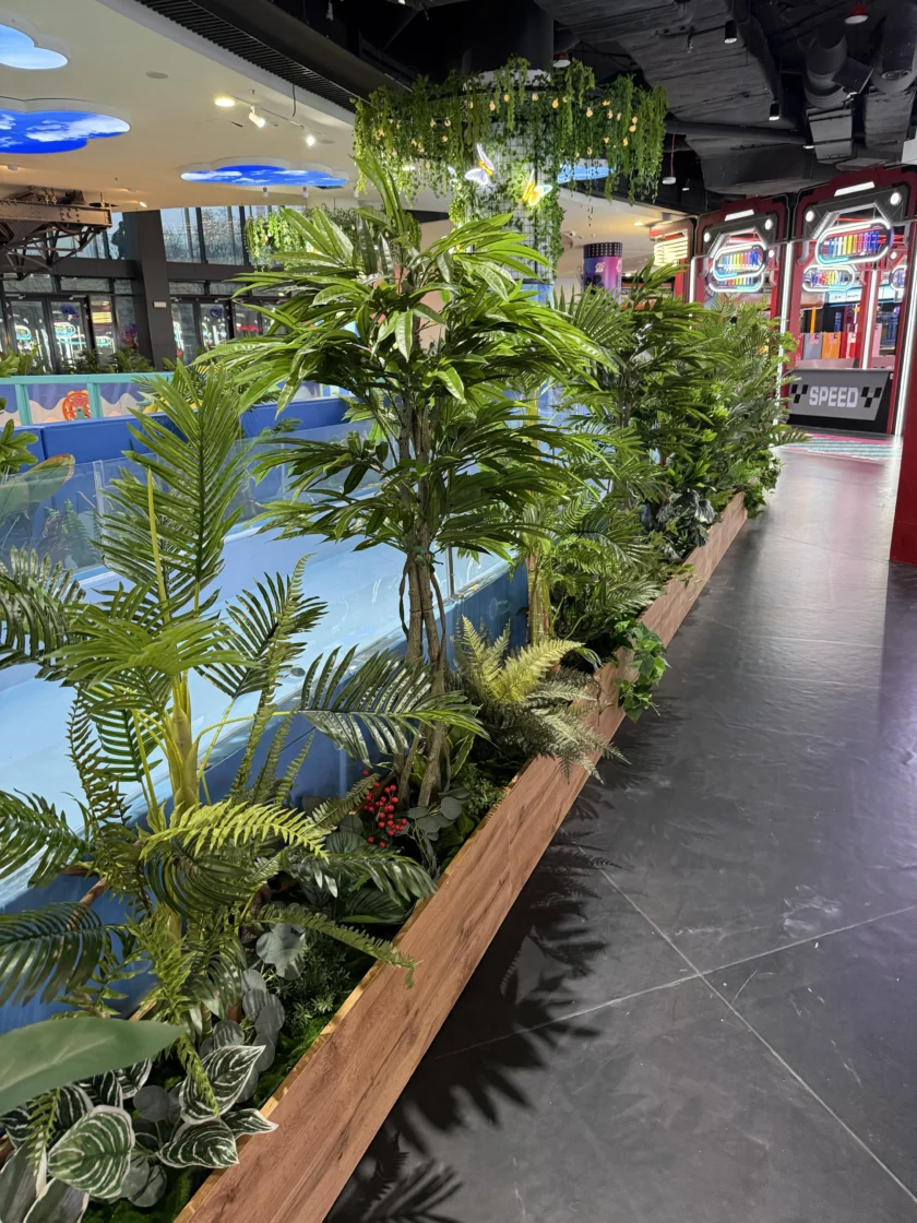 Kryty chodnik z drewnianą donicą wypełnioną zielonymi roślinami obok lodowiska, w tle automaty do gier.