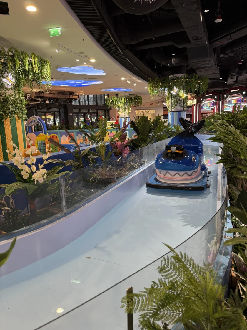 Niebieska łódź w kształcie rekina na torze wodnym otoczonym roślinami i sztuczną zielenią w krytej strefie rozrywki.