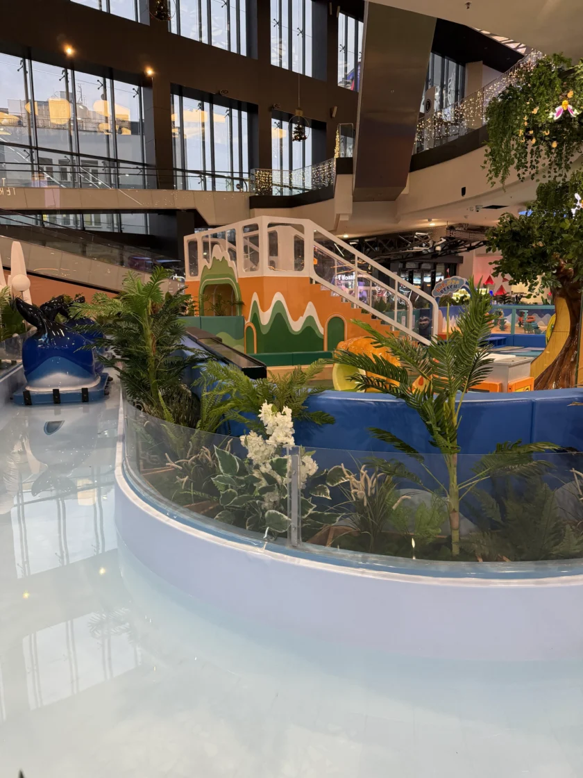 Kryty plac zabaw ze zjeżdżalnią zaprojektowaną tak, aby wyglądała jak góra, otoczony sztucznymi roślinami i szklanymi barierami, wewnątrz nowoczesnego budynku z dużymi oknami.