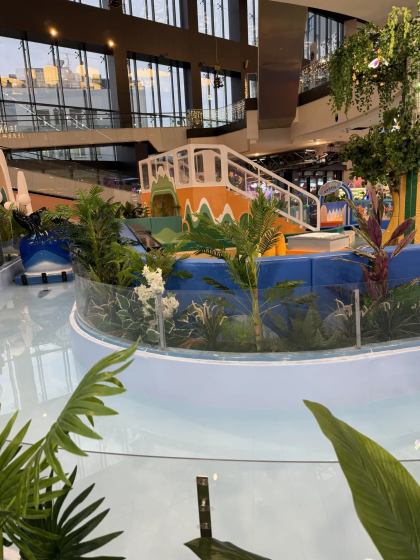 Kryty plac zabaw z kolorową zjeżdżalnią, sztucznymi roślinami, szklanymi barierkami i dekoracyjnym mostem, znajdujący się wewnątrz nowoczesnego centrum handlowego.