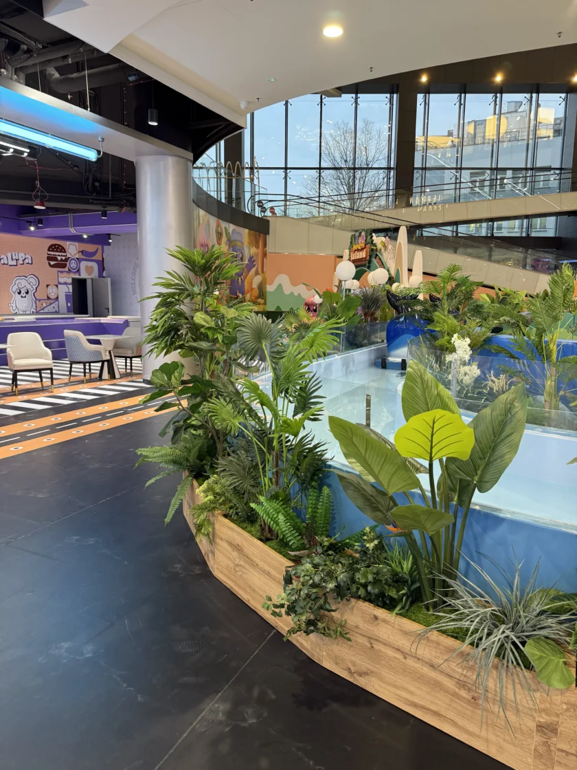 Kryty obszar centrum handlowego z okrągłym elementem wodnym z roślinami, pobliskimi miejscami do siedzenia i dużymi oknami z widokiem na budynki na zewnątrz.
