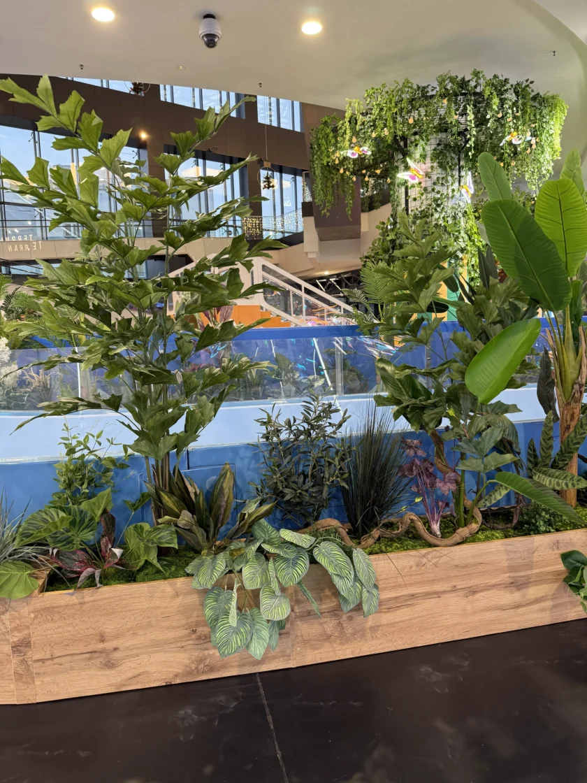 Sadzarka wewnętrzna z różnymi roślinami tropikalnymi przed atrakcją w postaci basenu z niebieską falą, ze schodami i zielenią wiszącą w tle.