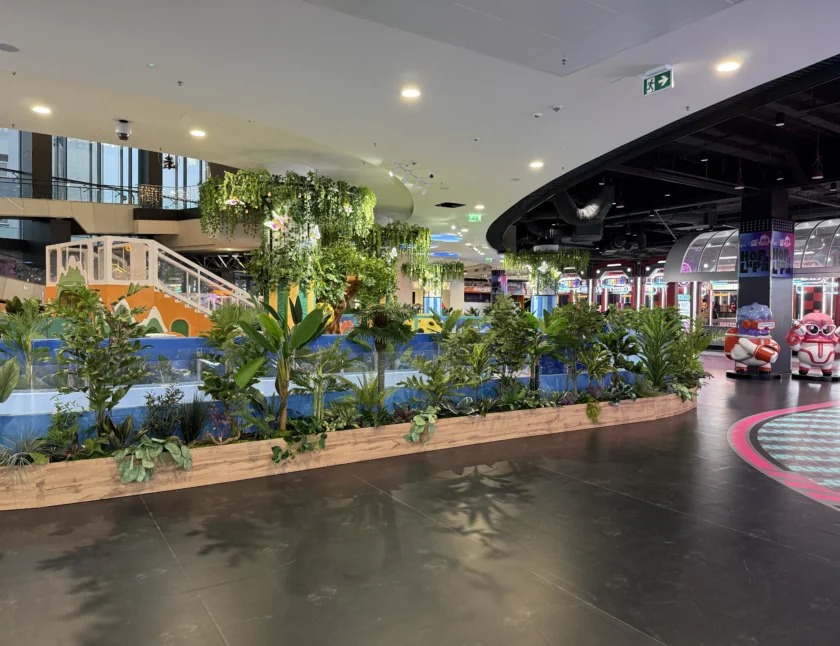 Kryta strefa rekreacyjna z ogrodem pełnym zielonych roślin, małym mostkiem i kolorowymi przejażdżkami w tle.