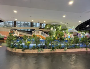 Wewnętrzny plac zabaw ze sztucznymi roślinami, małym białym mostkiem i kolorowym sprzętem do zabawy, ustawiony w przestronnym, dobrze oświetlonym otoczeniu.