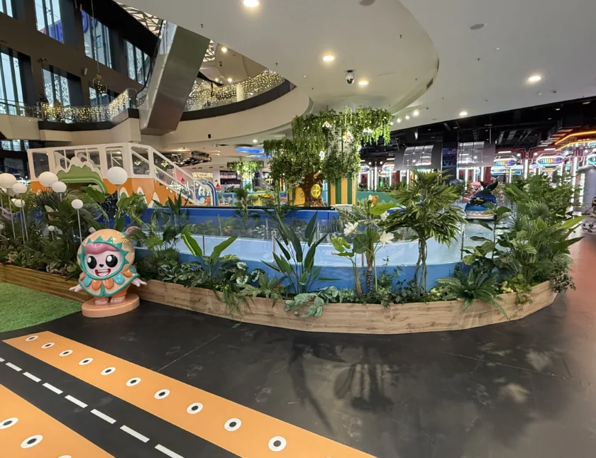 Kryty plac zabaw z małym basenem, sztuczną zielenią, ozdobnymi roślinami, zjeżdżalnią i posągiem postaci z kreskówek, w jasno oświetlonym centrum handlowym.