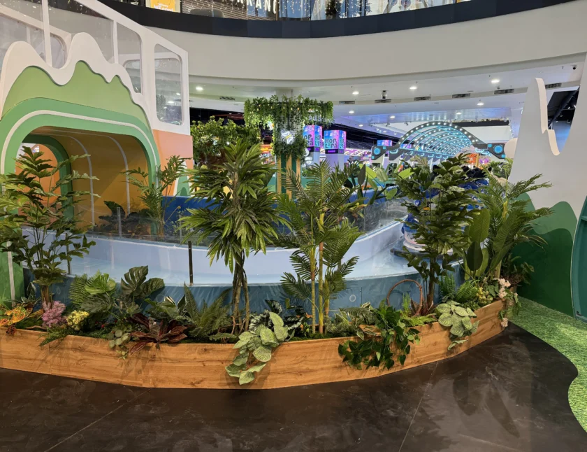 Wewnętrzna ekspozycja z drewnianą donicą wypełnioną różnymi zielonymi roślinami, zakrzywioną szklaną barierą i kolorowymi konstrukcjami tematycznymi w centrum handlowym.