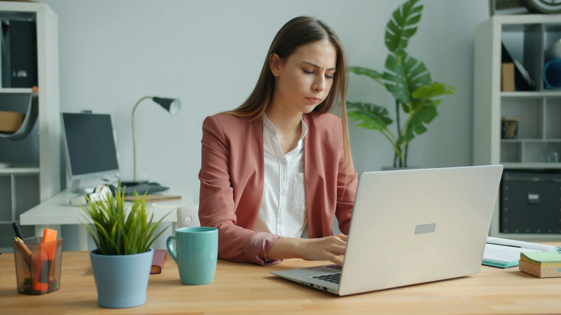Kobieta w różowej marynarce pracuje na laptopie przy drewnianym biurku, na którym stoją rośliny, niebieski kubek i artykuły biurowe, w nowoczesnym wnętrzu biurowym.