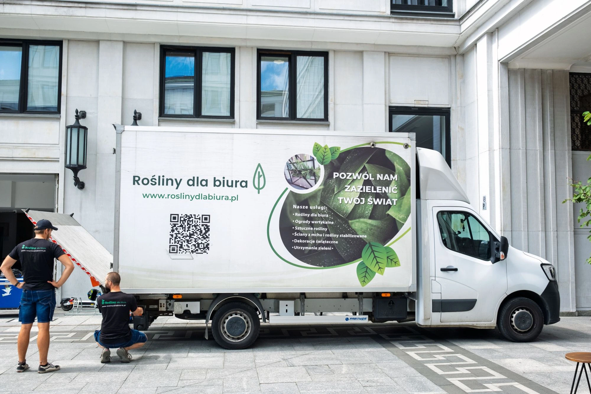 Dwóch pracowników rozładowuje białą furgonetkę dostawczą z napisem „Rośliny dla biura” i grafikami przedstawiającymi rośliny, zaparkowaną przed wejściem do nowoczesnego budynku.
