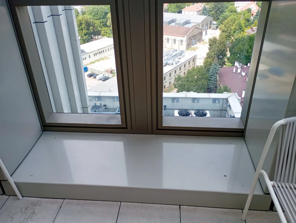 Duże okno z szerokim parapetem wychodzi na budynki, drzewa i dachy; w prawym dolnym rogu widać fragment białej ławki.