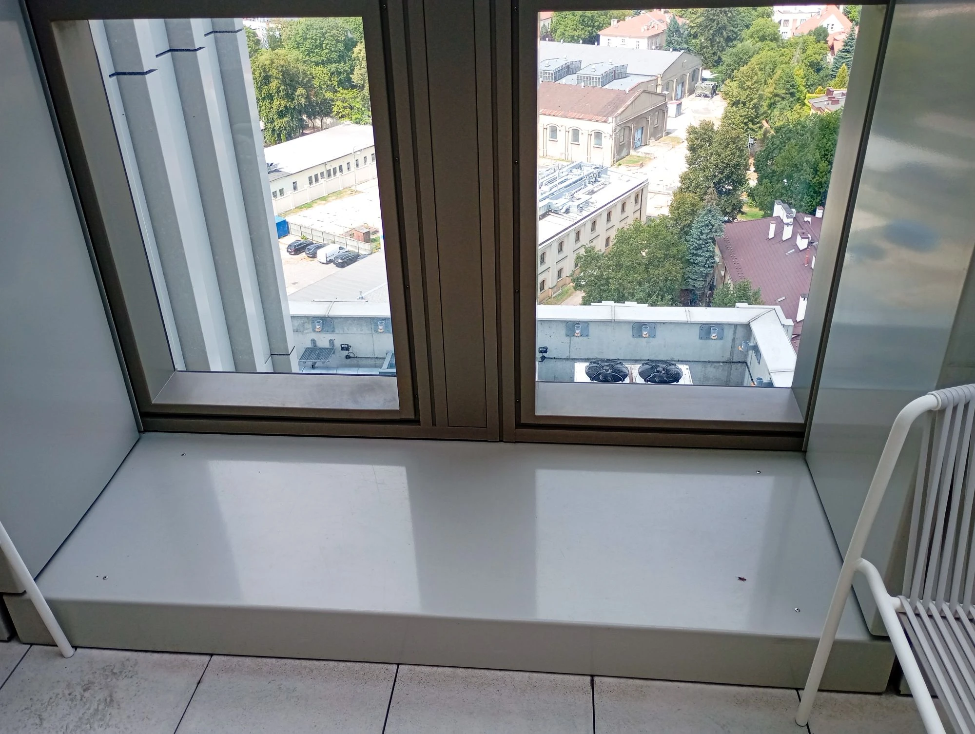 Duże okno z szerokim parapetem wychodzi na budynki, drzewa i dachy; w prawym dolnym rogu widać fragment białej ławki.