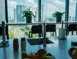 Z nowoczesnej wyspy kuchennej, na której stoją miski z owocami i różne przybory kuchenne, rozciąga się widok na panoramę miasta z wysokimi budynkami i roślinami w doniczkach na parapecie.