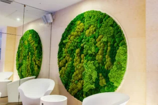 Okrągła instalacja ścienna wykonana z mchu w różnych odcieniach zieleni, której odbicie widoczne jest w dużym lustrze umieszczonym tuż obok, nad dwoma białymi krzesłami.